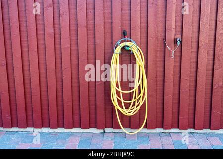 Yellow rubber watering tube for plants watering hangs on red wooden wall of traditional Swedish garden shed close to wall mounted water tap. Stock Photo