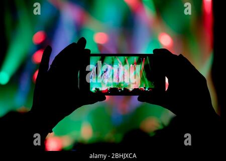 Collecting digital memory is loosing capability of being present, silhouette of a person hand shooting the concert colorful stage light effects with s Stock Photo