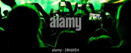 Collecting digital memory is loosing capability of being present, silhouettes of crowd of people shooting the concert with  mobile phones, in green li Stock Photo