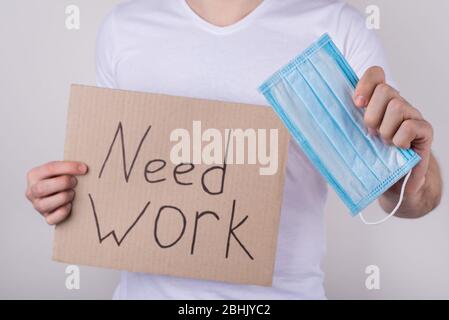 Economical crisis and quarantine concept. Cropped close up photo of guy holding  cardboard placard and surgical mask in hands isolated over grey backg Stock Photo