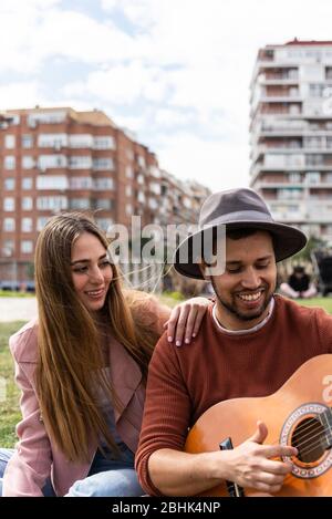 A young Hispanic man plays the guitar next to a young Caucasian woman in a city park Stock Photo