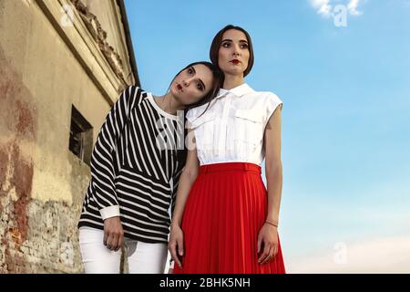 Outdoor fashion portrait of two young beautiful women wearing fashionable clothes posing on the street against the background of an old abandoned buil Stock Photo