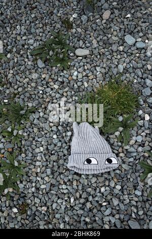 Knitted woollen grey child's beanie hat with eyes, abandoned & lying on ground surrounded by gravel & weeds, viewed from above Stock Photo