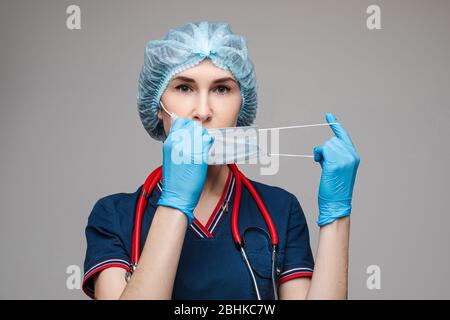 Handsome young caucasian female with long dark hair in dark blue t-shirt, a white gown, white trousers, medical hat with a phonendoscope wears a medic Stock Photo