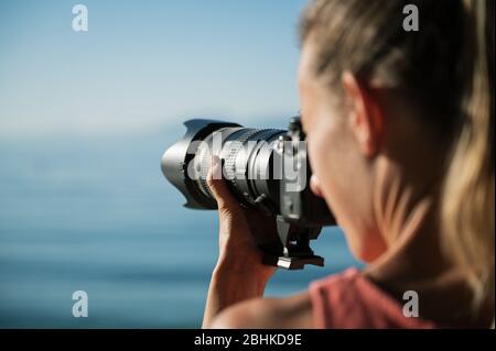 Over the shoulder view of female photographer taking photo of beautiful summer sea with professional dslr camera. Stock Photo