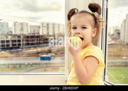 a little girl of 3-5 years old sits by the window and happily bites an apple. fruit in children's nutrition Stock Photo