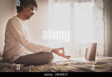 Mindful healthy man sitting on the bed in lotus position, practicing online yoga at home, connected with his laptop in quarantine, focus on his hand i Stock Photo