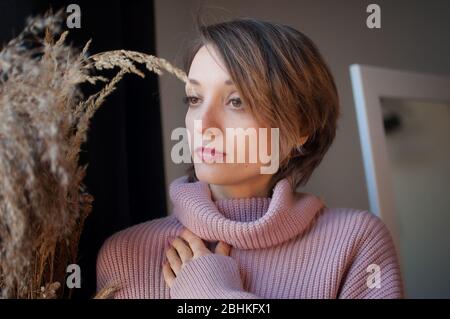 Attractive young woman looking at the bouquet of spikelets. Blonde girl with short hair wearing pink sweater in scandinavian interior. Atmospheric Stock Photo