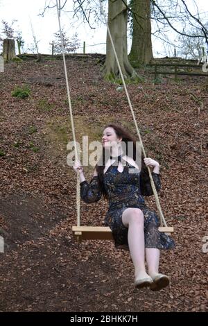 A long haired brunette young lady in a formal dress playing on a swing in winter woodland, smiling and happy Stock Photo