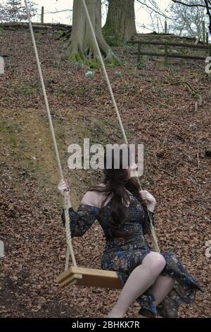 A long haired brunette young lady in a formal dress playing on a swing in winter woodland Stock Photo