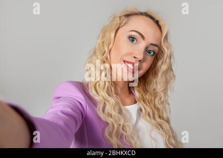 Closeup portrait of a stylish beautiful blonde woman with blue eyes, taking a selfie on a smartphone, video calling friends through an application, pleased with a smile, white background Stock Photo