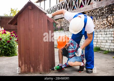 Cute boy in a construction helmet and glasses with a screwdriver in his hands building wooden dog house with his father. Construction and decoration Stock Photo