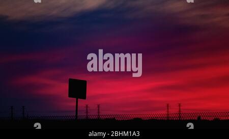 A road sign and electrical transmission towers form silhouettes against a vibrant pink, magenta and purple evening sky after sunset. Stock Photo