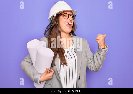 Young hispanic architect woman wearing builder hat and holding blueprints over yellow background pointing and showing with thumb up to the side with h Stock Photo