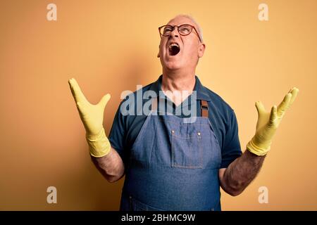 Middle age cleaner man cleaning wearing apron and gloves over isolated yellow background crazy and mad shouting and yelling with aggressive expression Stock Photo