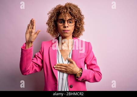 Young african american businesswoman wearing glasses standing over pink background Swearing with hand on chest and open palm, making a loyalty promise Stock Photo