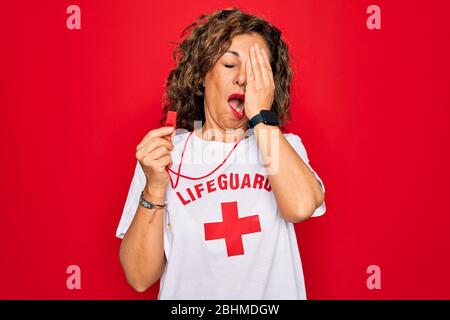 Middle age senior summer lifeguard woman holding whistle over red background Yawning tired covering half face, eye and mouth with hand. Face hurts in Stock Photo