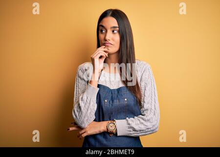Young beautiful baker woman wearing apron uniform cooking over yellow background with hand on chin thinking about question, pensive expression. Smilin Stock Photo