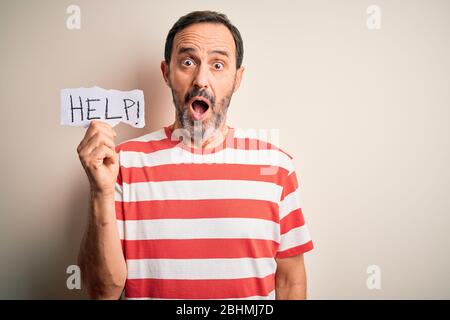 Middle age hoary man holding paper with help message over isolated white background scared in shock with a surprise face, afraid and excited with fear Stock Photo