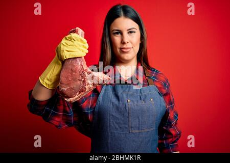Young butcher woman holding fresh raw beef meat stake over red background with a confident expression on smart face thinking serious Stock Photo