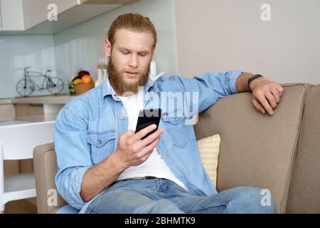 Serious young bearded man holding smartphone using online app on sofa at home. Stock Photo