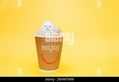 white chicken egg with a funny painted face in an eco-friendly paper Cup filled with paper on a bright yellow background. Stock Photo