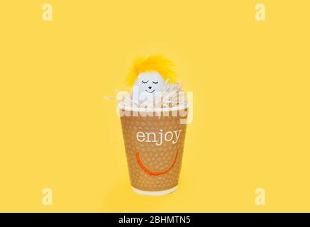 white chicken egg with a funny painted face and a crown of yellow feathers in an eco-friendly paper Cup filled with paper on a bright yellow backgroun Stock Photo