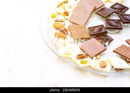 Set of assorted broken handmade chocolate bars pieces with nuts, dried berries, cranberries in a plate over white background. Overhead view, copy space. Stock Photo