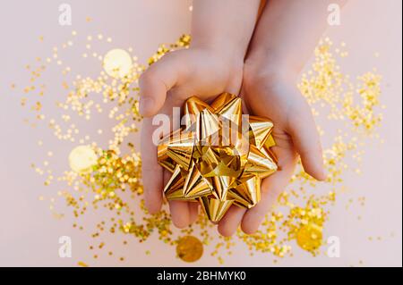 Children's hands light skin holding a large gift bow made of gold foil on a background of scattered gold sequins Stock Photo