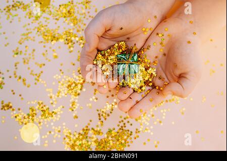 light-skinned hands hold a small green foil gift against a background of scattered gold stars on a soft beige background. Stock Photo