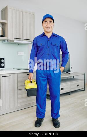 Smiling Asian handyman in blue uniform holding toolbox and smiling at camera Stock Photo