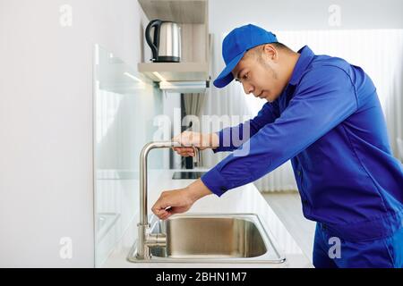Repairman in cap and uniform fixing water tap in kitchen sink Stock Photo