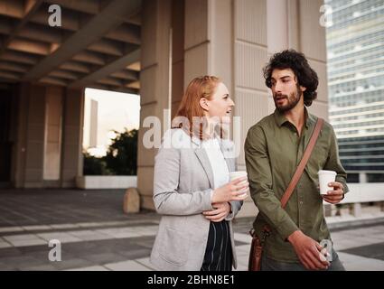 Male and female business partners drinking coffee in a conversation outdoor while walking out of building Stock Photo