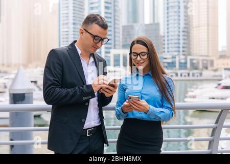 Two smiling business workers with mobile phone in hands are having pleasant conversation on skyscrapers background Stock Photo