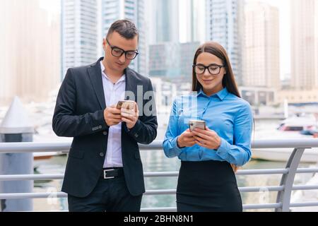 Two smiling business workers with mobile phone in hands are having pleasant conversation on skyscrapers background Stock Photo