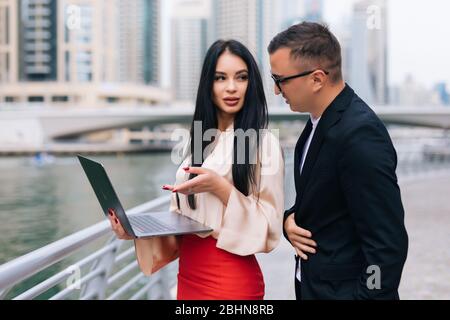 Two smiling young business people working on laptop and discuss something while sitting outdoors Stock Photo