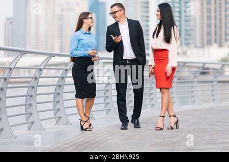 Excited business colleagues going along outdoor office glass wall. Business man and women walking outside, using tablet, drinking coffee, talking. Bus Stock Photo