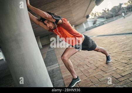 Portrait of a fitness man with armband and earphone doing stretching exercises under the bridge