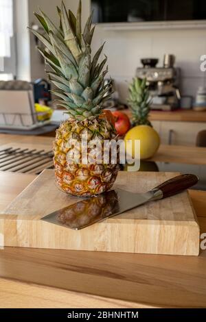 A cutting board with a whole pineapple fruit and a cleaver standing on a worktop in the kitchen in the background are more fruits and kitchen utensils Stock Photo