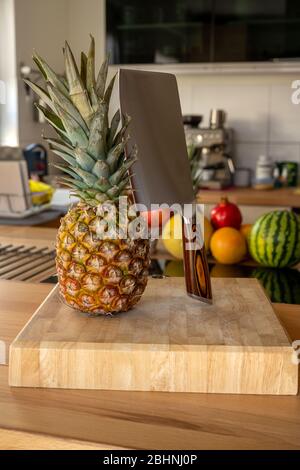 A cutting board with a whole pineapple fruit and a cleaver standing on a worktop in the kitchen in the background are more fruits and kitchen utensils Stock Photo