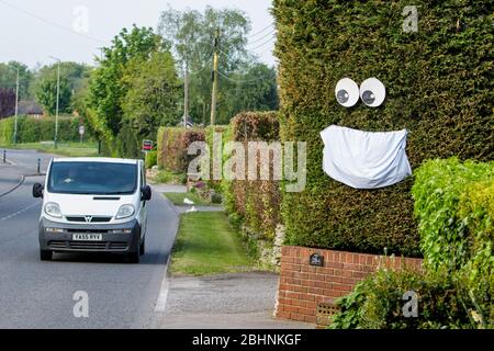 Chippenham, Wiltshire UK, 27th April, 2020. As scientific advisers in the UK discuss whether the public should wear face masks in public a van is pictured driving past a large face mask that has been hung on a hedge in Chippeneham, Wiltshire.  Credit: Lynchpics/Alamy Live News Stock Photo