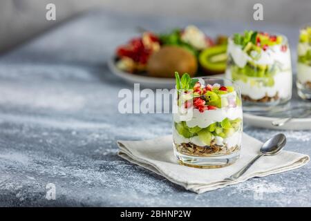 Glass jar of kiwi parfait dessert in glass with ingredients and spoon on gray napkin. Yogurt, granola and fruits. Healthy snack or breakfast. Light gr Stock Photo