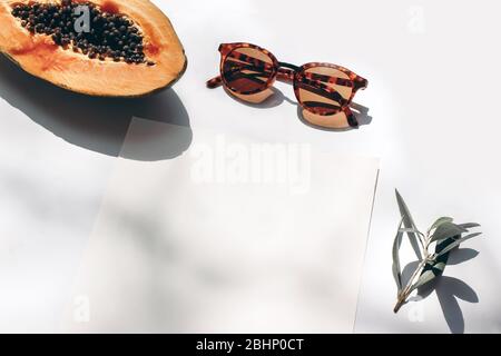 Summer stationery still life scene. Closeup of cut papaya fruit, olive branch and sunglasses in sunlight. White table. Blank paper cards, invitations Stock Photo