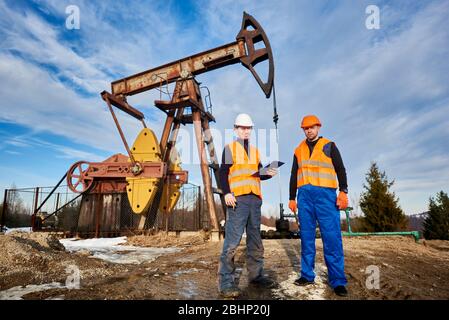 Two oil men in helmets and work vests standing on territory of oil field with oil pump rocker-machine and sky on background. Petroleum engineer holding clipboard, working with colleague in oil field. Stock Photo