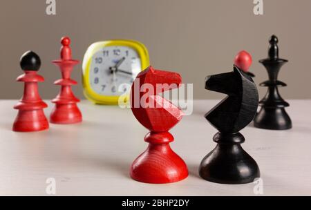 Two knights facing each other on a white wooden table, with an alarm clock and other chess pieces in the background Stock Photo