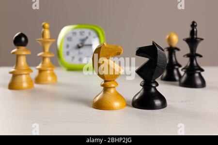 Two knights facing each other on a white wooden table, with an alarm clock and other chess pieces in the background Stock Photo