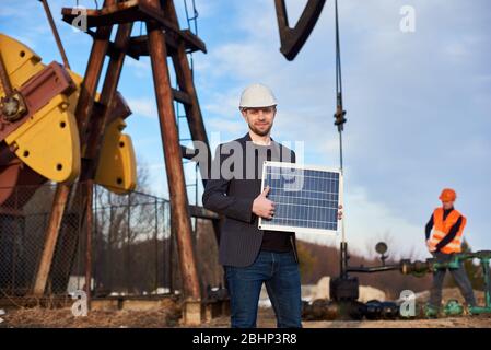 Portrait of smiling businessman wearing suit jacket and helmet, holding mini solar panel, showing thumb up. Oil operator working with pump jack on territory of oil field on blurred background. Stock Photo