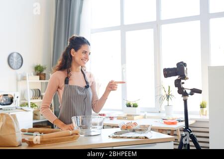Attractive young woman wearing apron standing in modern kitchen shooting video for her food blog Stock Photo