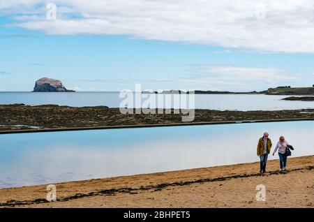 North Berwick, East Lothian, Scotland, United Kingdom, 27th April 2020. UK Weather: a calm bright day.  Despite the calm weather the beaches are almost empty during lockdown. The tidal bathing pool and the Firth of Forth are very still with the Bass Rock gannet colony on the horizon. A couple stroll on the beach in Milsey Bay Stock Photo