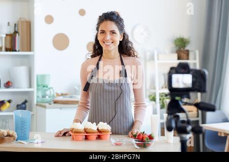 Portrait of young adult woman standing at kitchen table with homemade cupcakes with buttercream on it Stock Photo